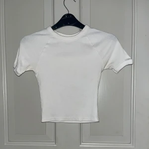 Vit ribbad topp H&M - Enkel och stilren vit topp från H&M med ribbad struktur. T-shirten har korta ärmar och rund halsringning, perfekt att matcha med jeans eller kjol för en clean look.