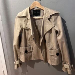 Beige trenchcoat från Zara Basic - Kort beige trenchcoat från Zara Basic med klassisk krage, stora svarta knappar och bältdetaljer vid ärmsluten. Jackan har två fickor framtill med lock och är tillverkad i ett bomullsliknande material. Perfekt för dig som gillar stilrena och tidlösa plagg.