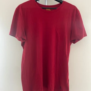 Röd Ralph Lauren T-shirt - Snygg röd t-shirt från Polo Ralph Lauren i klassisk modell med rund hals och korta ärmar. Tillverkad i mjuk bomull och har den ikoniska broderade loggan i svart nedtill. Perfekt för en clean och stilren look.