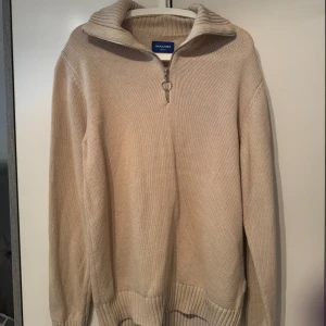 Beige stickad half zip från Jack & Jones - Beige stickad tröja från Jack & Jones med ribbad hög krage och snygg half zip-dragkedja med ringdetalj. Långärmad och i mjukt material som passar perfekt när det är kyligt. Enkel och stilren design som är lätt att matcha.