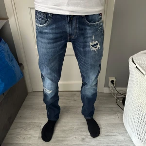 Blå Replay jeans med slitningar - Säljer ett par blå Replay jeans med snygga slitningar och trasiga detaljer på låret och knät. Jeansen har raka ben, klassiska fem fickor och tydliga kontrastsömmar. Märkespatch bak i midjan och logga vid fickan. Perfekta för en avslappnad streetstil.