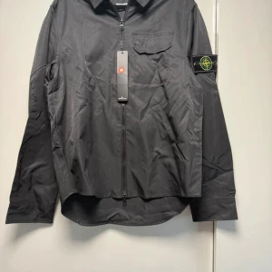Svart overshirt från Stone Island - Svart overshirt från Stone Island med klassisk krage, dragkedja framtill och bröstficka. Ikonisk logga på vänster ärm. Tillverkad i lätt polyester, perfekt för lager-på-lager. Snygg och clean look som funkar till det mesta.