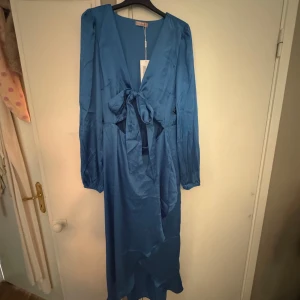 Blå midiklänning med knyt framtill - Supersnygg blå långklänning från Nelly i glansigt material. Klänningen har v-ringning, långa ärmar och ett brett knytband i bröstet med en cutout som ger en fin siluett. Perfekt för dig som vill sticka ut med en elegant look. Färgen stämmer överens med bild 1 och 2.