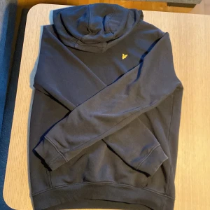 Svart hoodie från Lyle & Scott Junior - Svart hoodie från Lyle & Scott Junior med klassisk huva och gul broderad logga på bröstet. Mjuk insida och ribbade muddar vid ärmslut och nederkant. Perfekt för en chill och stilren look.