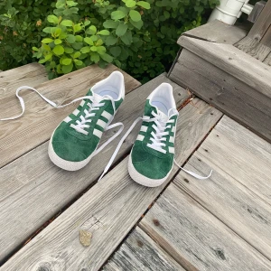 Adidas Gazelle gröna sneakers stl 36 - Säljer ett par gröna Adidas Gazelle sneakers i mocka med vita detaljer och klassiska tre ränder på sidorna. Skorna har vit sula, snörning och guldtext på sidan. Perfekta för dig som gillar retrostil och vill sticka ut med färg.