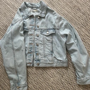 Ljusblå jeansjacka från Gina Tricot - Säljer en klassisk ljusblå jeansjacka från Gina Tricot i storlek M. Jackan har knappar framtill, bröstficka med lock och knapp samt snygga sömdetaljer. 