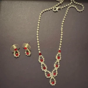 Glittrigt halsband och örhängen med röda stenar - Set med halsband och örhängen i silverfärg med gnistrande klara stenar och röda kristaller. Halsbandet har droppformade detaljer och örhängena matchar med samma form och färg