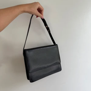 Svart axelväska i skinn - Stilren svart axelväska i slätt skinn med justerbart axelband och enkel, boxig form. Väskan har ett fack med lock framtill och en dragkedjeficka på baksidan. Perfekt för dig som gillar minimalistisk och klassisk design.