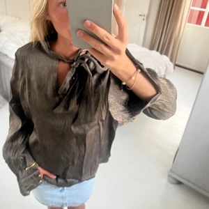 Brun glansig blus med knyt i halsen - Säljer en brun glansig blus i ett lätt skimrande material. Blusen har långa ärmar med breda manschetter och guldiga knappar. Snygg knytning i halsen och avslappnad passform.