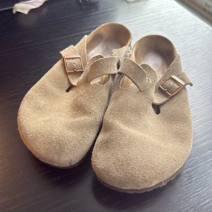 Birkenstock Boston - Beige mocka - Snygga sandaler från Birkenstock i beige mocka med rund tå och justerbart spänne i metall. Skorna har en platt sula och klassisk slip-in design, perfekt för en avslappnad stil. Mjukt material och bekväm fotbädd för vardagskomfort.