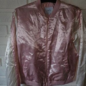 Rosa satin bomberjacka från NEU - Snygg bomberjacka i glansigt satinmaterial från NEU. Jackan är ljusrosa med vita ärmar och har en rosa rand längs ärmarna. Stängs med dragkedja framtill och har ribbade muddar vid hals, ärmslut och nederkant. Perfekt statement-plagg för dig som gillar att sticka ut.