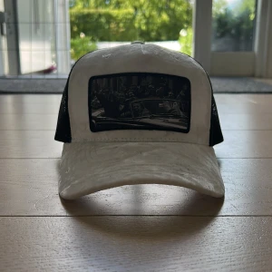 Vit och svart truckerkeps från Hatstore - Snygg truckerkeps i vitt och svart från Hatstore. Framsidan har ett svartvitt tryck med motiv av en bilscen, och baksidan är i svart mesh för extra ventilation. Justerbar passform med klassisk böjd skärm. Perfekt för en cool och avslappnad stil.