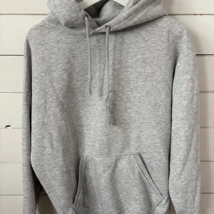 Grå hoodie från bikbok  - En stilren ljusgrå hoodie med huva och snörning, samt en magficka framtill. Tröjan har lång ärm och är tillverkad i bomull och polyester. Perfekt för chill dagar eller när du vill ha en enkel och clean look.