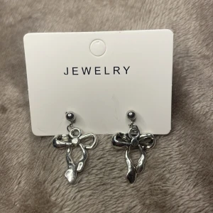 Silvriga örhängen med rosett - Snygga silvriga örhängen med hängande rosettform. De har en blank finish och en liten detalj i mitten av rosetten. Perfekta för att ge din look en söt och trendig touch.