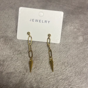 Guldiga kedjeörhängen med spetsig detalj - Snygga örhängen i guldton med kedjelänkar och en cool, spetsig konformad detalj längst ner. Perfekta för dig som vill ha något trendigt och edgy i öronen. Lätta att matcha med andra smycken och ger en modern vibe till din stil.