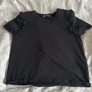 Svart volangtopp från ZARA - Svart topp från ZARA med korta ärmar och snygga volangdetaljer på axlarna. Den är i storlek S men skulle säga att den sitter mer som en xs. väldigt enkel och snygg💘