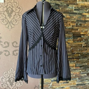 Svart randig skjorta med spetsdetaljer - Svart skjorta med vita ränder och snygga spetsdetaljer vid ärmslut och över bröstet. Långärmad modell med knappar framtill och klassisk krage. Tillverkad i lätt, stretchig polyesterblandning. S enligt stl, men passar nog mer en M