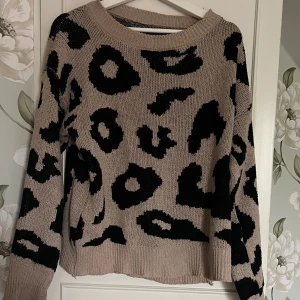 Beige leopardmönstrad stickad tröja - Säljer en beige stickad tröja med svart leopardmönster. Tröjan har en rund halsringning, långa ärmar och ribbade muddar. 