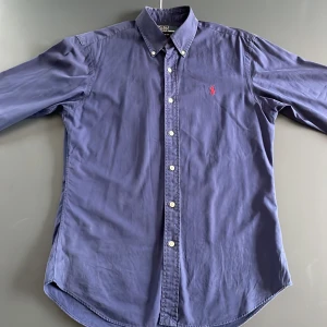 Marinblå skjorta Polo Ralph Lauren M - Snygg marinblå skjorta från Polo Ralph Lauren i slim fit-modell. Skjortan har klassisk krage, vita knappar och rött broderat logotyp på bröstet. Tillverkad i bomull och har lång ärm. Perfekt för dig som gillar stilrena och tidlösa plagg.