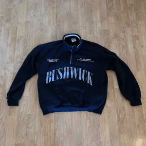 Svart half zip tröja från Bershka - Svart sweatshirt från Bershka med stor BUSHWICK-print framtill och vit text på bröstet och ryggen. Tröjan har half zip, ståkrage med ribbad kant och långa ärmar med muddar. 