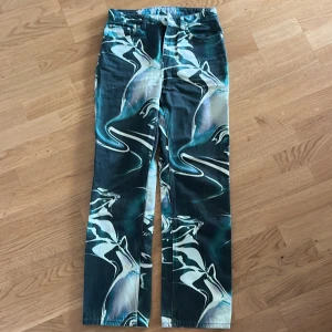 Weekday Voyager swirl-mönstrade jeans - Säljer ett par unika Weekday Voyager jeans med swirl-mönster i grönt, blått och vitt. Byxorna har rak passform och normal midja. Materialet är kraftig bomull som ger en snygg och cool look. Perfekta för dig som vill sticka ut med din stil.