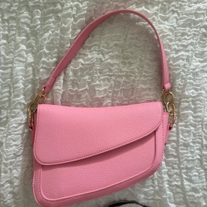 Rosa axelväska i skinnimitation - Säljer en snygg rosa axelväska med guldiga metalldetaljer och stilren design. Helt oanvänd 