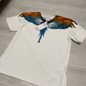 Marcelo Burlon  - (Passar folk har har storlek M också) Säljer en vit Marcelo Burlon t shirt med ikoniskt vingtryck i orange, blått och svart över axlar och bröst. T-shirten har rund halsringning och korta ärmar. Materialet är mjuk bomull och passformen är normal. Perfekt statement-plagg för dig som gillar unika prints.