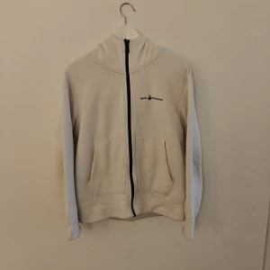 Sail racing kofta - Vit kofta med dragkedja från Sail Racing. Modellen har huva, två stora fickor fram och svart logga på bröstet samt på huvan. Tillverkad i mjuk bomull och polyester, perfekt för chill dagar. Snygg och enkel stil som funkar till det mesta.