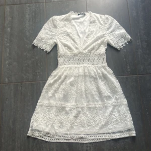Vit spetsklänning från Gina Tricot - Superfin vit kortklänning från Gina Tricot med v-ringning, spetsdetaljer och markerad midja. Klänningen har korta ärmar med spetskant och vackert broderat tyg. 