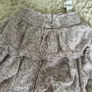 Blommig volangkjol från Stradivarius XS - Supersöt kort volangkjol från Stradivarius i ljuslila med små vita och rosa blommor samt gröna blad. Kjolen har smock i midjan och flera lager volanger som ger en flowy känsla. Perfekt för vår och sommar!