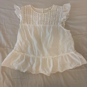 Vit blus med spets och volang H&M - Supersöt vit blus från H&M med spetsdetaljer framtill och volang vid ärmslut och nederkant. Blusen är ärmlös med små volangärmar. 