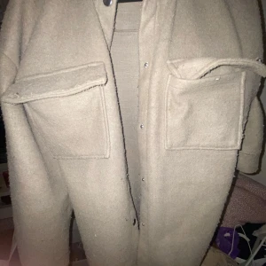 Beige overshirt i ull med stora fickor - Overshirt i beige ull med två stora bröstfickor och tryckknappar framtill. Jackan har en loose passform och lång ärm, perfekt för lager-på-lager. Enkel och stilren design som funkar till många outfits.