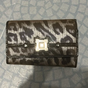 Leopardmönstrad plånbok med metallspänne - Snygg plånbok i svart och grått med leopardmönster och ett coolt metallspänne framtill. Flera fack för kort, mynt och sedlar. Materialet är glansigt och känns som skinn. Perfekt för dig som vill ha något unikt och trendigt i väskan.