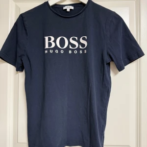Mörkblå BOSS t-shirt med logga - Snygg mörkblå t-shirt från Hugo Boss med stor vit logga framtill. Klassisk rund halsringning och korta ärmar. Tillverkad i mjuk bomull som känns skön mot huden. Perfekt för dig som gillar stilrena och trendiga plagg.