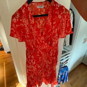 Röd blommig omlottklänning Soaked in Luxury - Superfin röd kortklänning från Soaked in Luxury med vitt blommönster. Omlottmodell med knyt i midjan, v-ringning och korta ärmar. Perfekt för sommarens alla tillfällen. Lätt och luftig känsla!