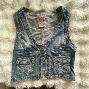 Blå jeansväst från Street One - Snygg blå jeansväst från Street One med klassisk v-ringning och dekorativa knappar framtill. Västen har två fickor med lock och coola sömdetaljer. Perfekt att slänga över en t-shirt för en avslappnad look.