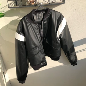 En svart skinnjacka i kortare/croppad passform - Snygg svart bomberjacka i skinnmaterial från Boohoo med vita detaljer på ärmarna. Jackan har dragkedja framtill, två stora fickor och ribbade muddar vid hals, ärmslut och nederkant. Den är oversize och croppad passform.