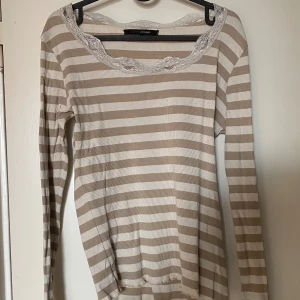 Randig långärmad topp med spets - Superfin långärmad topp från Tippy Concepts i beige och vitt med horisontella ränder. Söt spetsdetalj runt den breda ringningen och vid ärmsluten. Mjuk ribbad bomullskänsla, perfekt för lager-på-lager och chill vibes. Frakt tillkommer 
