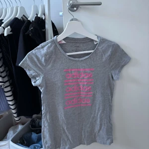 Grå Adidas t-shirt med rosa tryck - Snygg grå t-shirt från Adidas med korta ärmar och rund hals. Framsidan har ett coolt rosa Adidas-tryck i flera rader. Perfekt för en sportig och avslappnad stil. Mjuk och skön bomullskänsla.