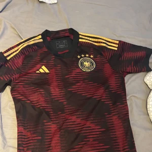 Tysklands matchtröja Adidas S - Säljer en officiell Tyskland fotbollströja från Adidas i storlek S. Tröjan är svart med röda och gula detaljer, har korta ärmar och ett unikt mönster. Tysklands landslagsemblem med fyra stjärnor på bröstet och klassiska Adidas-ränder på axlarna.