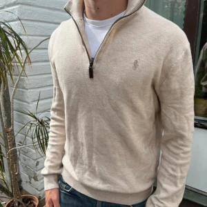Beige half zip tröja Polo Ralph Lauren - Snygg beige långärmad tröja från Polo Ralph Lauren i bomull. Klassisk modell med half zip och diskret broderad logga på bröstet. Perfekt till jeans eller chinos för en clean och stilren look.