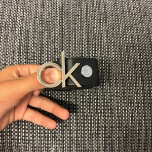 Svart skärp från Calvin Klein i skinn - Snyggt svart bälte från Calvin Klein i äkta skinn med stor silvrig CK-logga som spänne. Bältet är stilrent och har en clean look med diskret logga även på sidan. Perfekt accessoar för att lyfta din outfit.