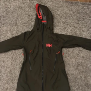 Svart regnjacka från Helly Hansen - Snygg svart regnjacka från Helly Hansen med röda detaljer och logga på bröstet och ryggen. Jackan har huva, hel dragkedja framtill och två stora fickor med dragkedja. Insidan är grå och materialet är syntet, perfekt för blöta dagar.