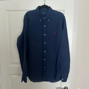 Marinblå skjorta från Ralph Lauren - Snygg marinblå skjorta från Ralph Lauren i slim fit-modell. Skjortan har klassisk krage, vita knappar och den ikoniska röda broderade loggan på bröstet. Tillverkad i bomull och har långa ärmar. Perfekt för dig som gillar stilrena och tidlösa plagg. Sjukt fräsch inför sommaren! 