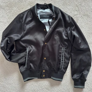 Svart bomberjacka från J.Lindeberg - Snygg svart bomberjacka från J.Lindeberg med ribbade muddar och krage med vita ränder. Jackan har tryckknappar i guld och två fickor framtill med randiga detaljer. Perfekt för dig som vill ha en stilren och sportig look.