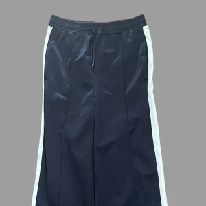 Svarta vida byxor med vita ränder - 🌟Snygga svarta byxor med vita ränder längs sidorna och bred passform. Byxorna har resår i midjan med snörning och är tillverkade i ett glansigt syntetmaterial. Perfekta för en sportig och avslappnad stil. Skickas snabbt📦