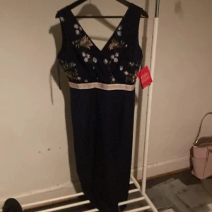 Mörkblå långklänning från Little Mistress - Supersnygg mörkblå långklänning från Little Mistress med v-ringning och broderade blommor upptill. Klänningen har en ljus midjerem och är ärmlös, perfekt för dig som vill sticka ut med elegans. Säljes med tag kvar!