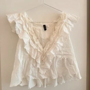 Vit volangblus från hm - Supersöt vit blus från hm med volanger längs v-ringningen och axlarna. Luftig och flowy modell i bomull, perfekt för en romantisk look. Ärmlös design och dubbla lager nertill för extra volym.