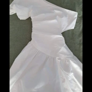 Vit oneshoulder kortklänning - Snygg vit kortklänning med oneshoulder-design och lätt rynkad midja. Kjolen är utsvängd och tyget känns luftigt, perfekt för varma dagar. Klänningen har en modern och stilren look som sticker ut.