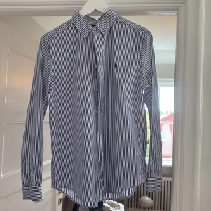 Randig skjorta från Ralph Lauren - Klassisk skjorta från Ralph Lauren med blåvita ränder och broderad logga på bröstet. Skjortan har lång ärm, knappar framtill och en snygg krage. Perfekt för dig som gillar stilrena och tidlösa plagg.